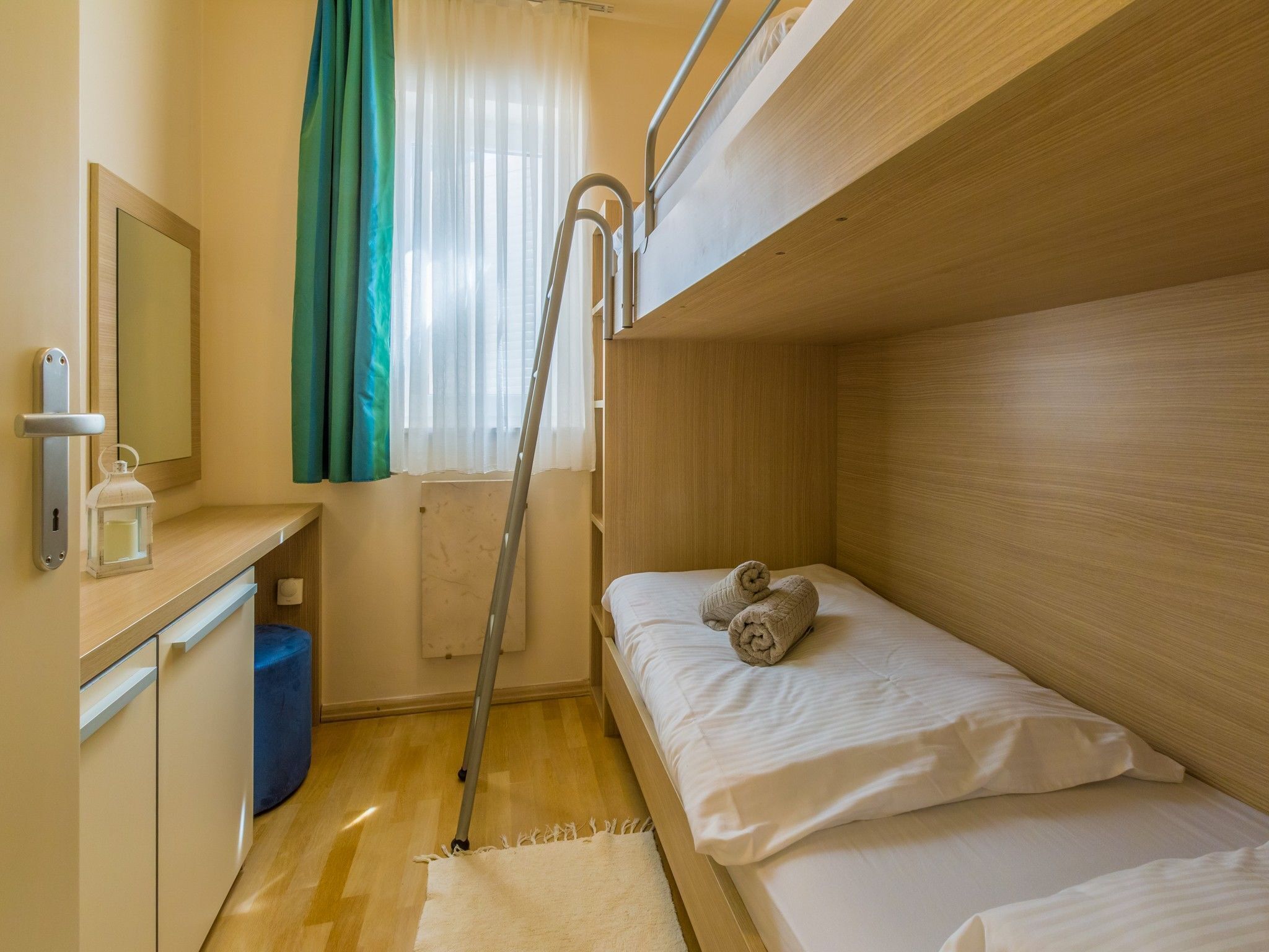 Ein kleines Zimmer mit einem Etagenbett, einem kleinen Schreibtisch mit Spiegel, einer Laterne und einem fenster mit Vorhängen.