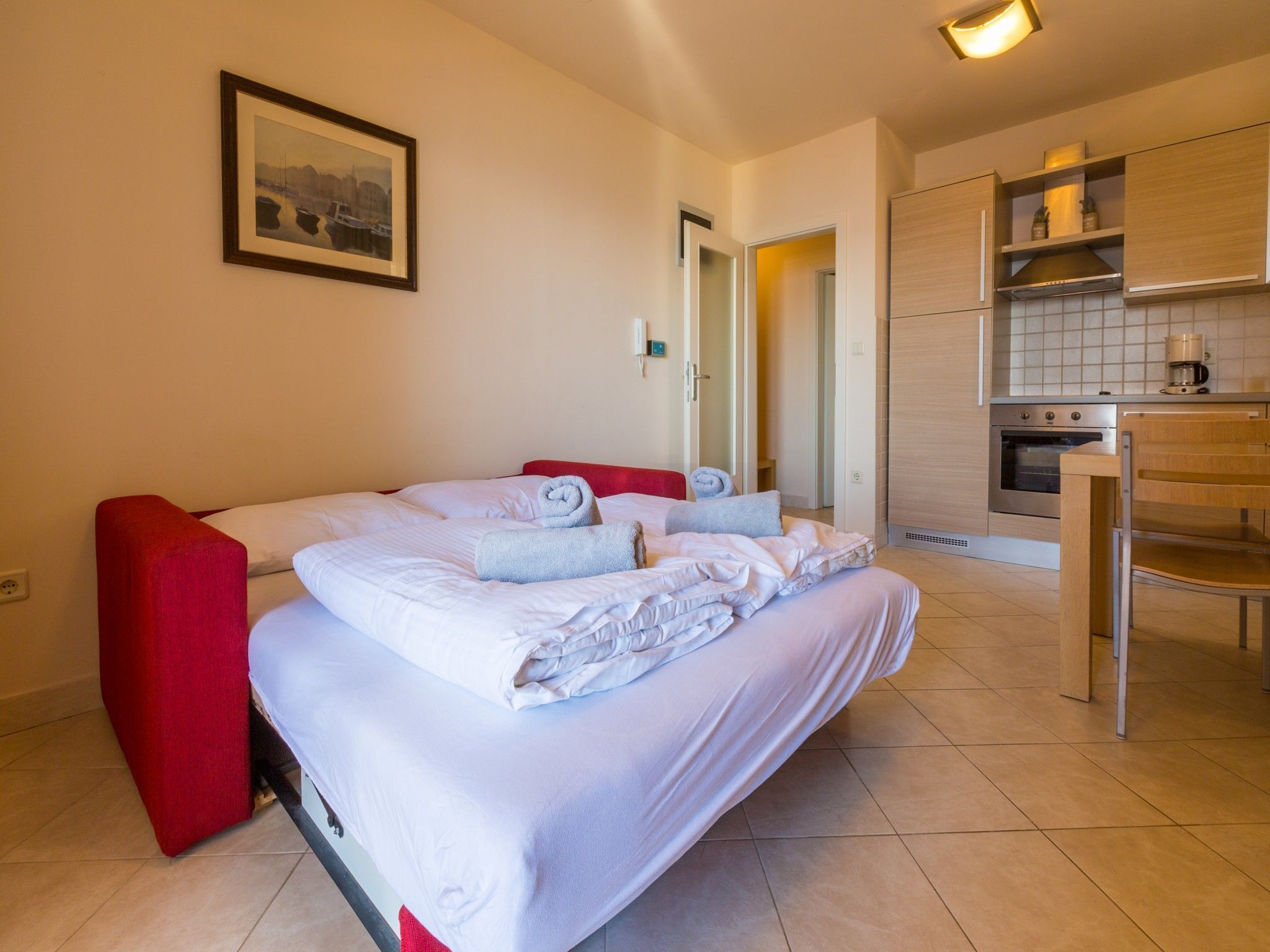A hotel room with a bed, a red chair, a kitchen area with a stove and a coffee maker, and a framed picture on the wall.