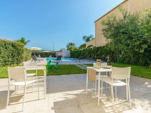 Zona piscina con sedie a sdraio e un tavolo, circondata da vegetazione e un edificio.