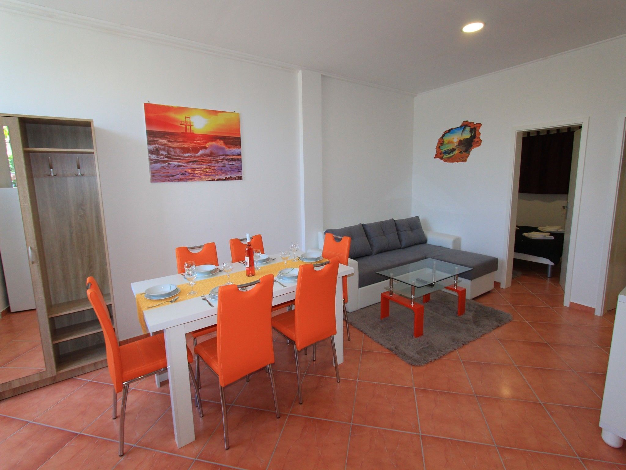 A modern living room with a dining area, featuring a sofa, orange chairs, a glass coffee table, and a painting of a sunset.