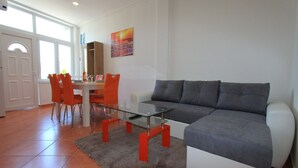 Ein modernes Wohnzimmer mit Glastisch, grauem Sofa, orangenen Sesseln und einem Essbereich mit weißem Tisch und orangenen Stühlen.