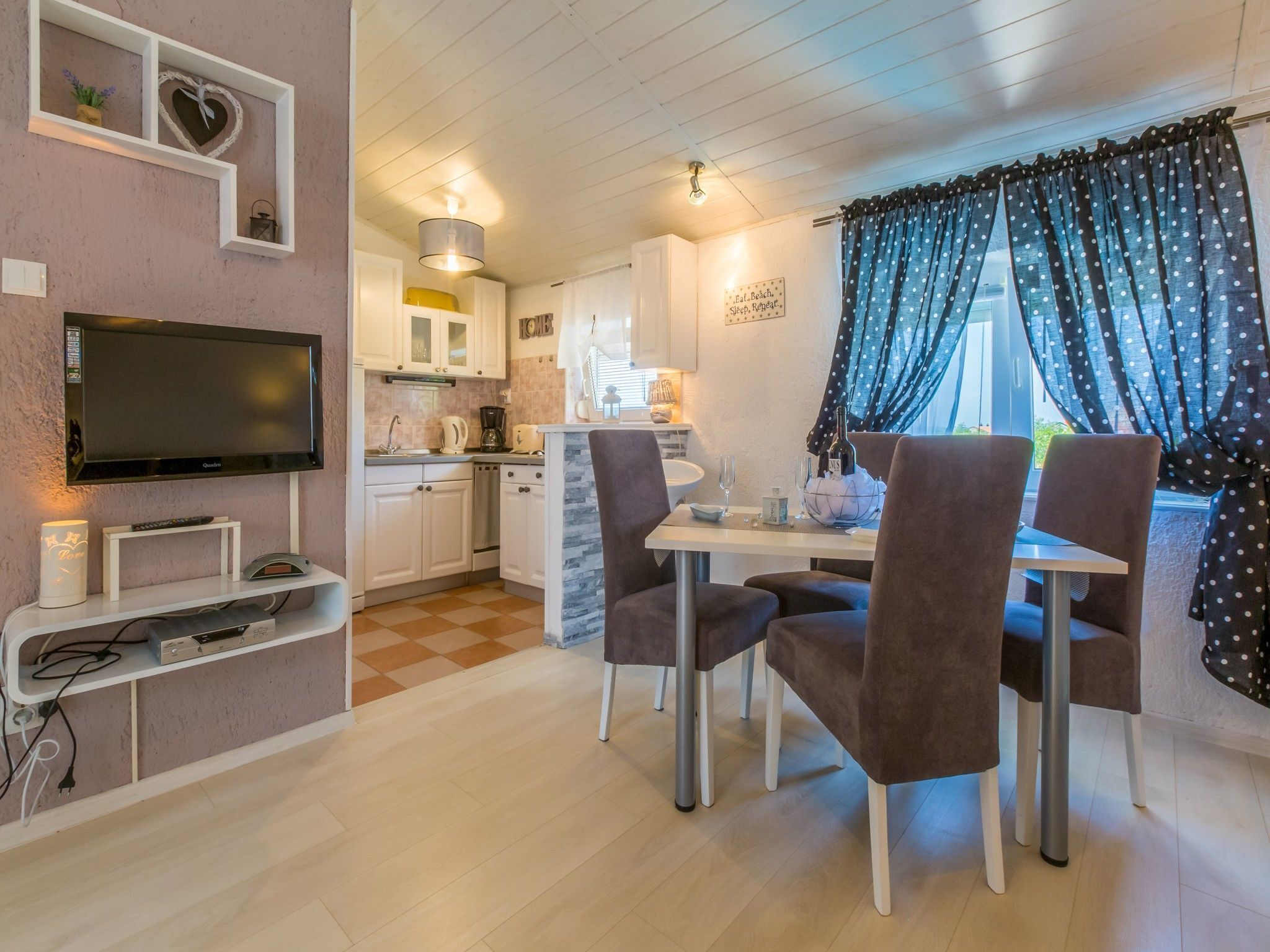 A modern kitchen with white cabinets, a built-in oven, and a flat-screen TV. There is a dining area with a table and chairs, and a window with polka dot curtains.