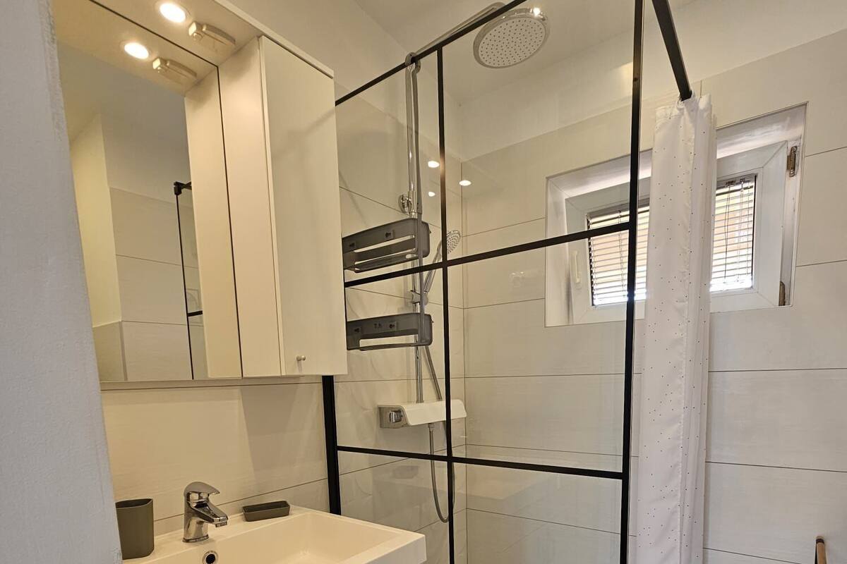A modern bathroom with a glass shower enclosure, a white sink with a mirror above, and a window with blinds.