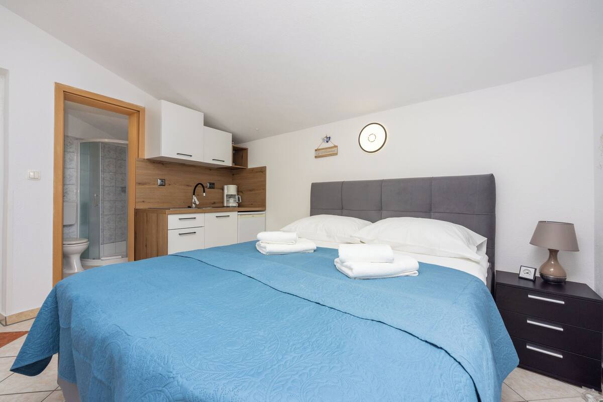 A bedroom with a bed, bedside table, and a view of the bathroom and kitchen.