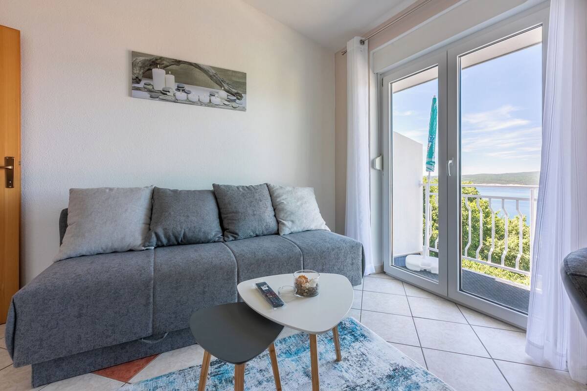 A modern living room with a grey sofa, a small table with a remote control, and a view of a balcony with a railing.