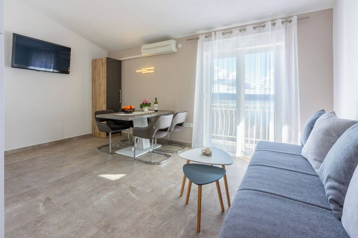 A modern living room with a grey sofa, a dining table with chairs, a flat-screen TV mounted on the wall, and a balcony with a view.