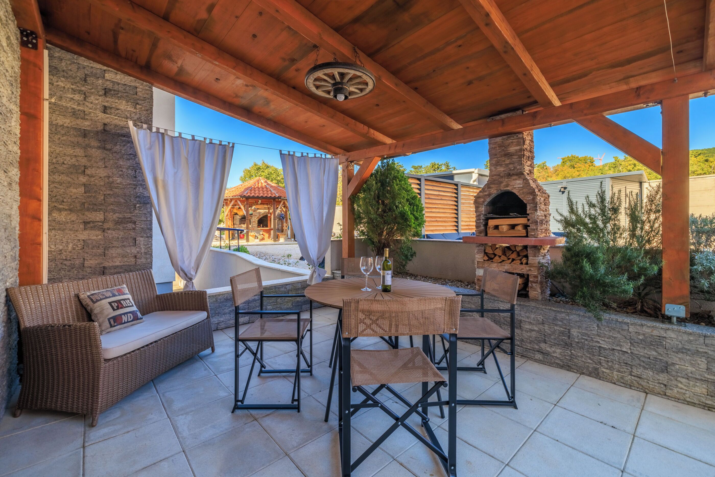 A covered patio with a wicker sofa, dining table, chairs, and a stone fireplace.