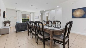 A dining area with a glass-top table surrounded by dark wooden chairs, white walls, and a large window with curtains. - 8BR Superb Villa, Pool, Spa • Near Disney (Kissimmee)