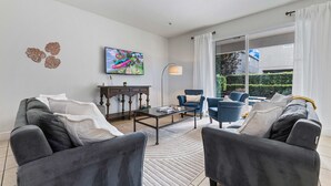 A modern living room with a grey sofa set, a coffee table, and a TV mounted on the wall. There is a large window with curtains, and a small table with a lamp. - 8BR Superb Villa, Pool, Spa • Near Disney (Kissimmee)