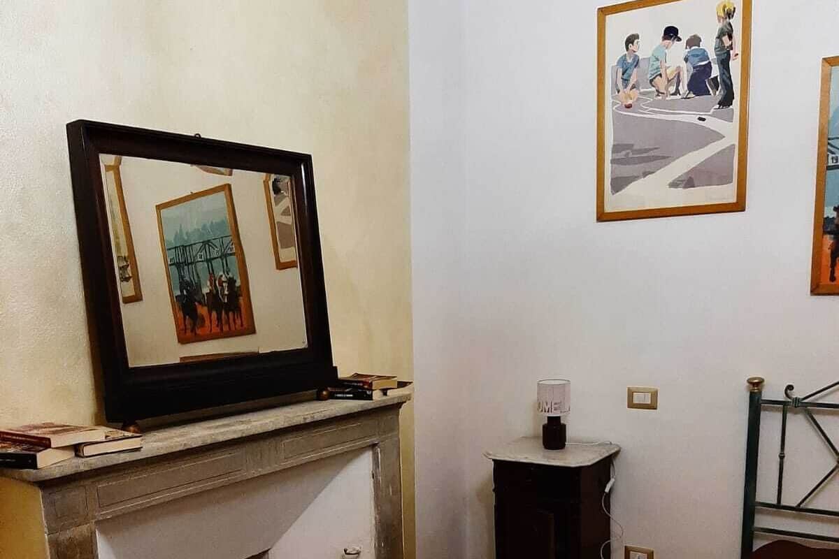 A room with a fireplace, a framed painting above it, a small table with a lamp, and a staircase with a metal railing.