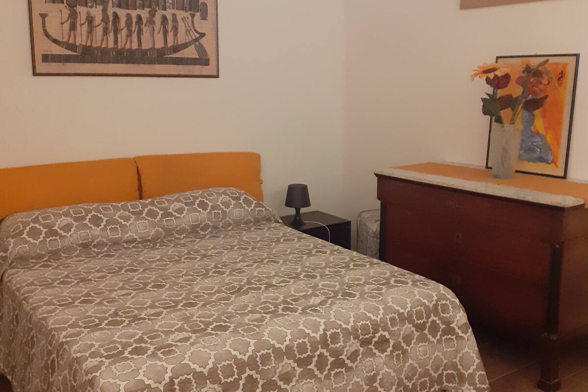 A bedroom with a patterned bedspread, a wooden nightstand, a desk with a vase of flowers, and two framed artworks on the walls.