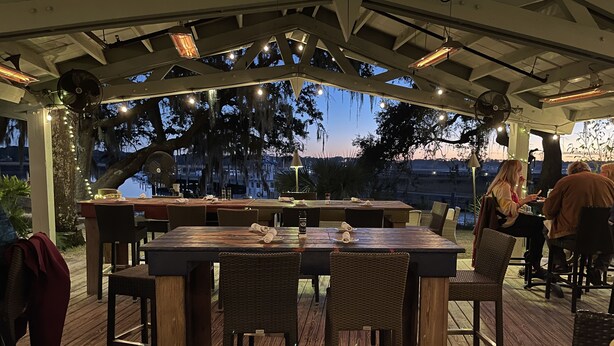 An outdoor dining area with a covered patio, wooden tables and chairs, string lights, and a view of trees and a body of water. - 8/27- 9/4  open  5 STARS  Great ratings- Hidden Gem Great Lagoon/Golf/pool view (Hilton Head Island)