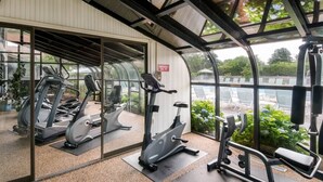 Une salle de sport équipée de matériel d’exercice, notamment des tapis de course et des appareils de musculation, et offrant une vue sur un espace extérieur aménagé avec des sièges et de la verdure, grâce à de grandes fenêtres.