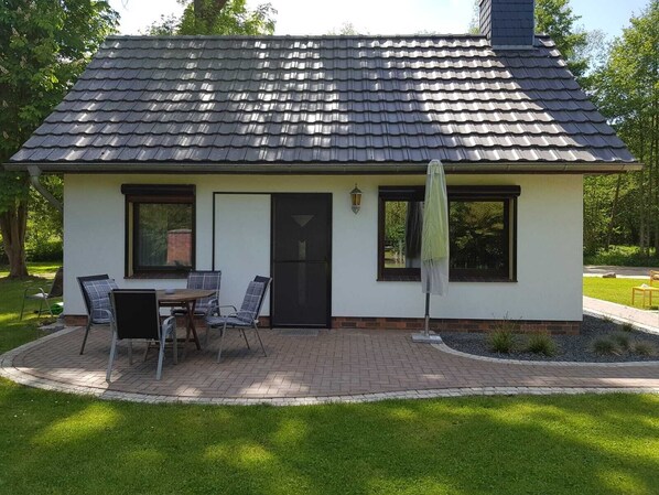 Une petite maison de plain-pied, avec un toit en tuiles, une terrasse en briques et une table avec des chaises à l’extérieur.