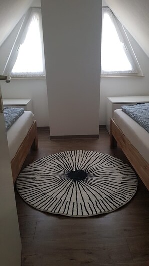 Une petite pièce bien éclairée, avec un tapis rond à motifs zébrés, deux lits en bois et un plafond blanc.