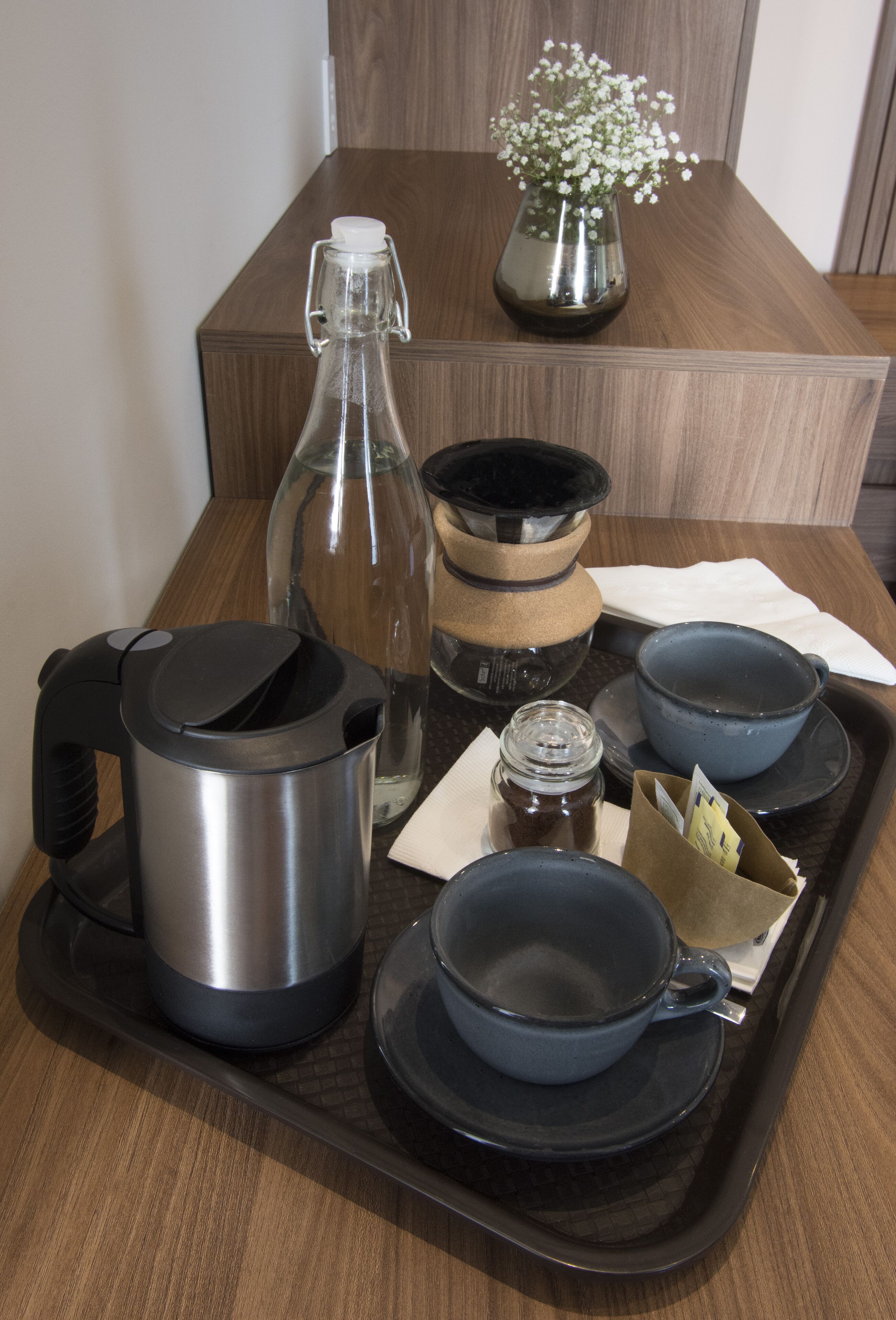 Un plateau contenant une bouilloire en acier inoxydable, une bouteille en verre, un bol bleu, un bol noir et un petit pot de confiture.