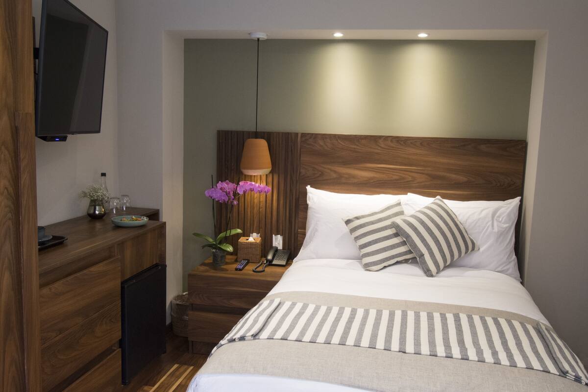 Une chambre à coucher bien rangée, avec une tête de lit en bois, une table de chevet avec une lampe, un téléviseur fixé au mur et un lit avec une literie à rayures.
