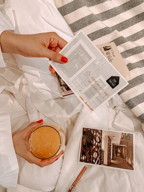Une personne tient une brochure sur laquelle on peut voir une photo d’un bâtiment, un verre de jus d’orange et un stylo, le tout posé sur un lit recouvert d’une couverture à rayures.