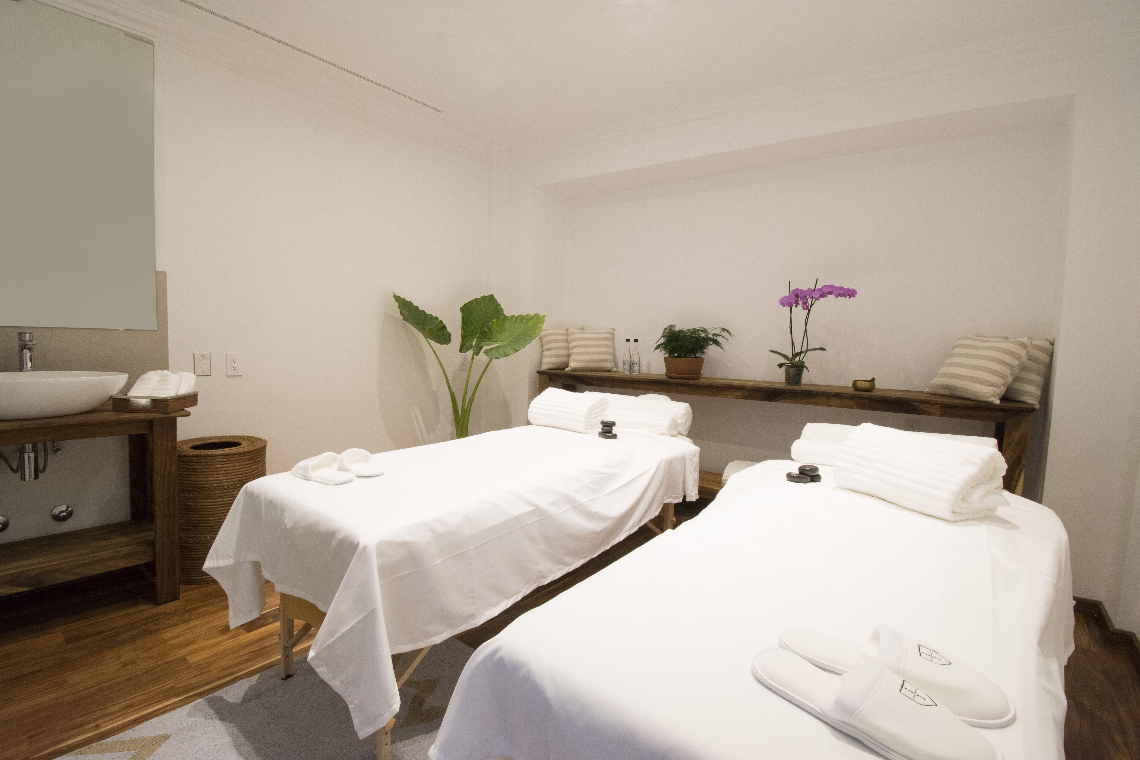 A clean, minimalist room with two beds, a large mirror, a wooden vanity, and a small shelf with decorative items.