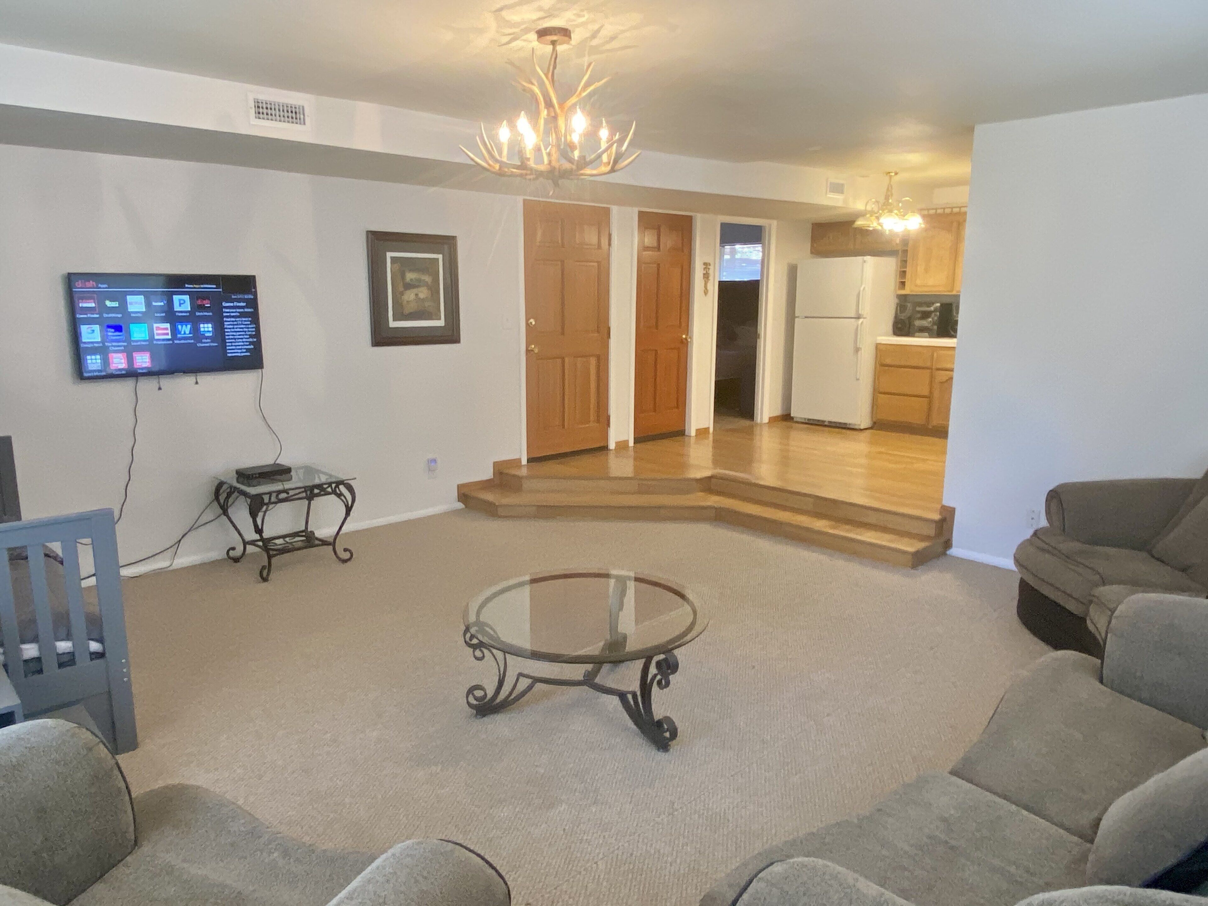 A living room with a flat-screen TV, a glass coffee table, and a sofa. There is a wooden door leading to a kitchen.