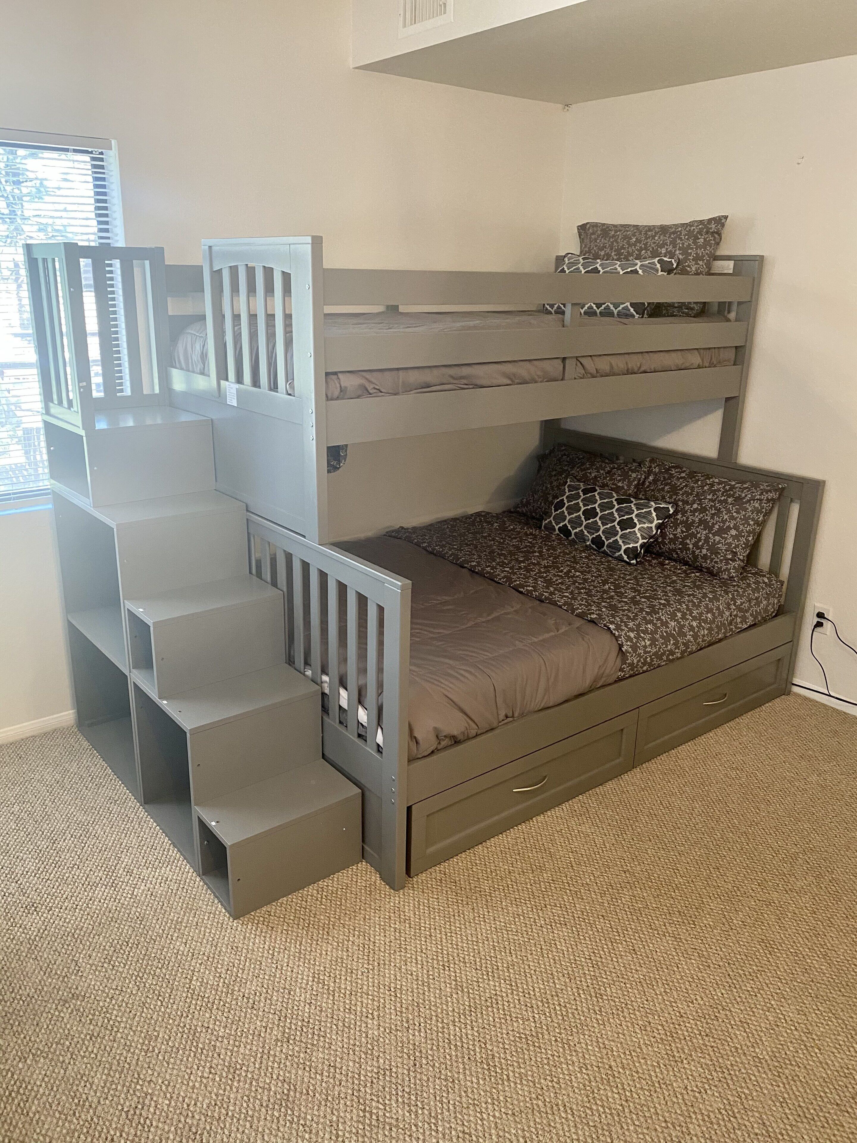 A bunk bed with a built-in desk and shelving, carpeted floor, and a window with blinds.