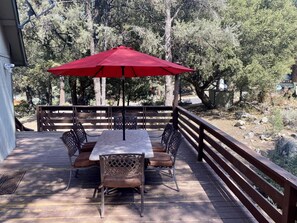 A wooden deck with a table and chairs under a red umbrella, surrounded by trees. - Quail Run 4,000 Sq.Ft Largest and Most Beautiful Cabin On The Mountain (Pine Mountain Club)