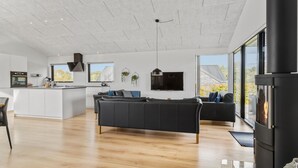 A modern living room with a fireplace, a sofa, a TV, and a kitchen area with a large window. - 4 bedroom cozy home in Haderslev (Haderslev)