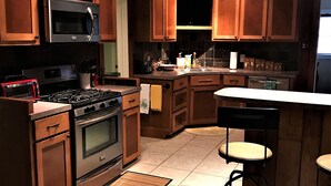 A kitchen with wooden cabinets, stainless steel appliances, a bar area with stools, and a ceiling fan. - Hughes Street House! (Nacogdoches)