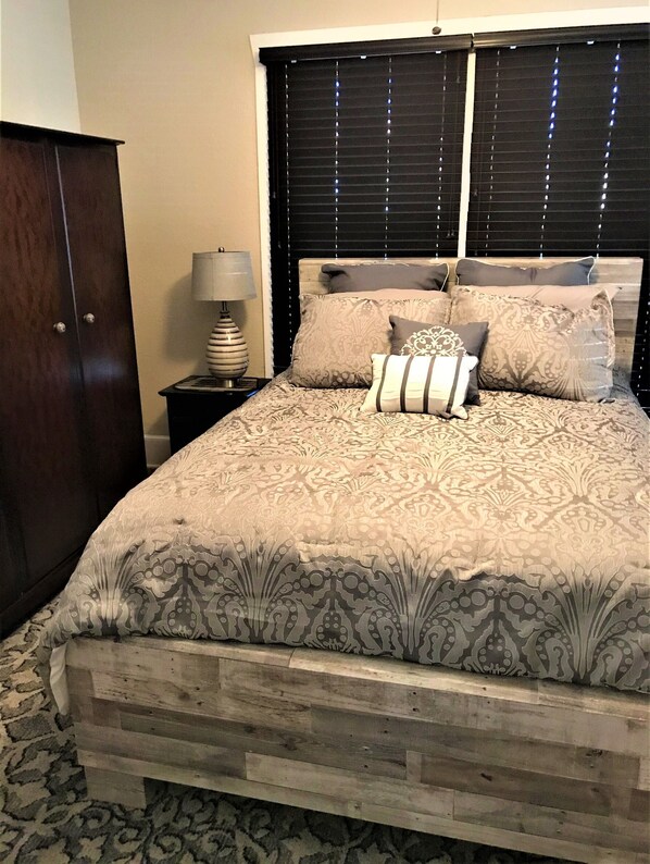 A bedroom with a wooden bed frame, patterned bedding, a nightstand with a lamp, and a dark-colored wardrobe. - Hughes Street House! (Nacogdoches)