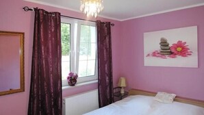 A bedroom with a bed, a window with curtains, a radiator, a painting on the wall, and a small table with a lamp.