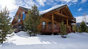 Una cabaña de troncos con techo nevado, rodeada de nieve y árboles.