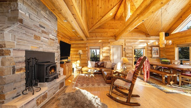 Interior de una cabaña de troncos con chimenea de piedra, vigas de madera, un rincón de descanso acogedor y una mesa de comedor.