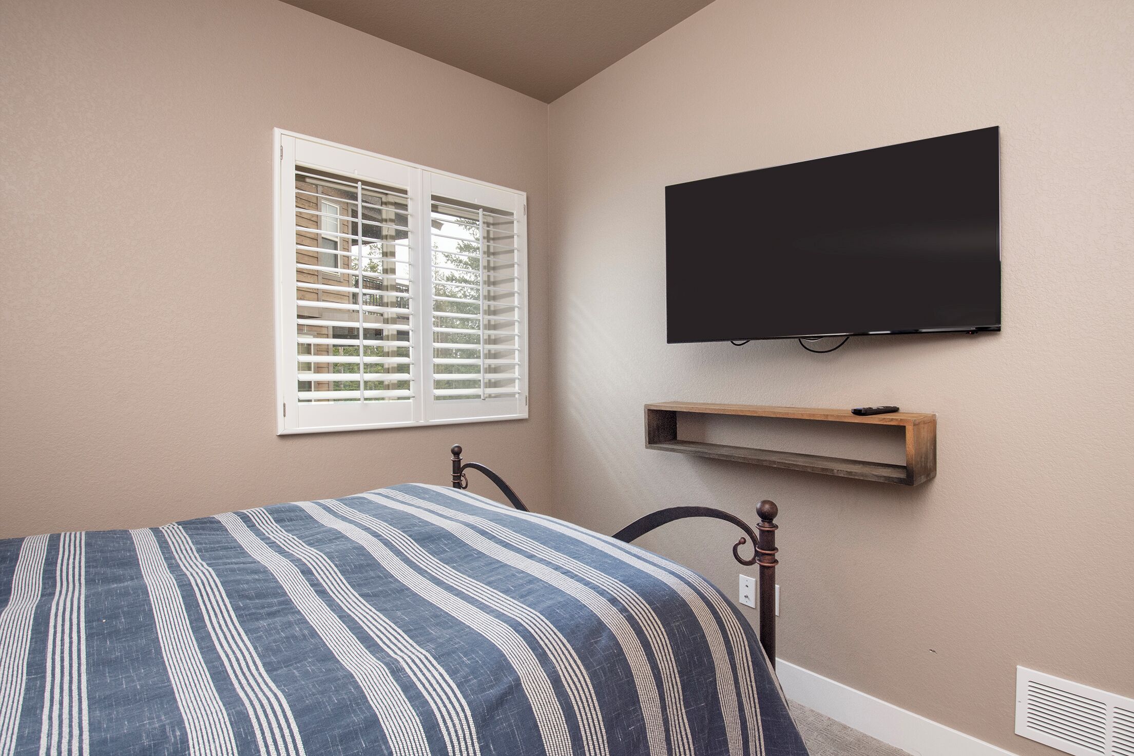 Un dormitorio con cama, ventana con persianas, televisor montado y estante.