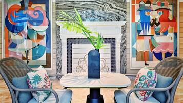 A room with a round table, two wicker chairs, a vase with green plants, and abstract paintings on the walls.