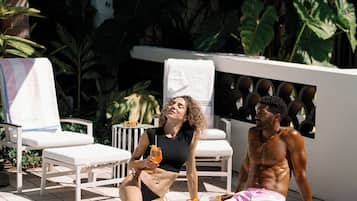 Un área junto a la piscina con sillones de descanso, una piscina con baldosas y plantas tropicales.