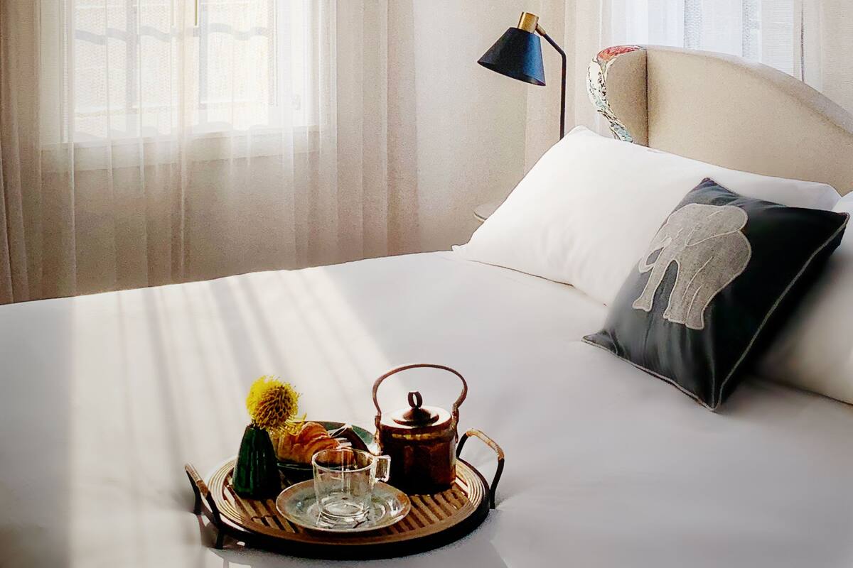 A bedroom with a bed, a bedside table with a lamp, a tray with a teapot, and sheer curtains.