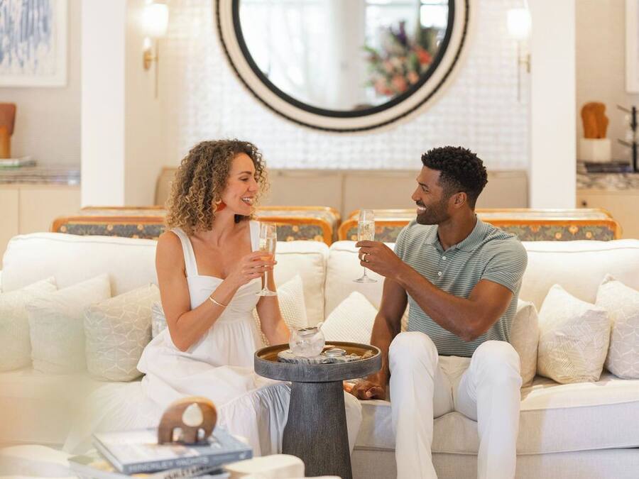 Un couple est assis sur un canapé blanc, dans une pièce bien éclairée. Un miroir rond et une petite table ronde se trouvent entre eux.