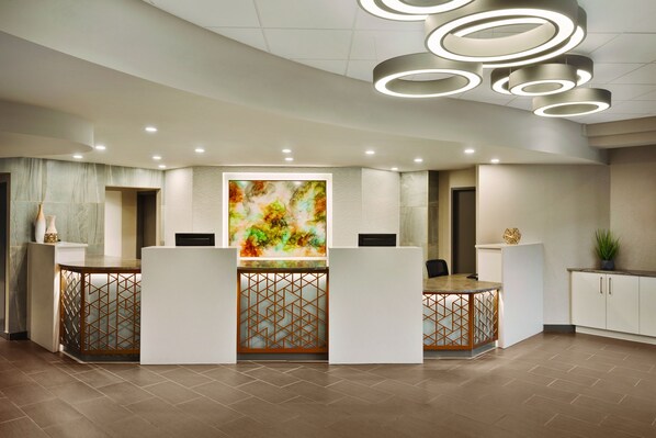Un área de recepción con diseño moderno, que presenta un mostrador central con patrón geométrico, un cuadro abstracto de gran tamaño y luminarias contemporáneas.