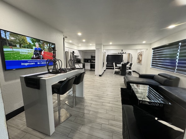 A modern living room with a TV, bar area, seating, and a kitchen in the background.