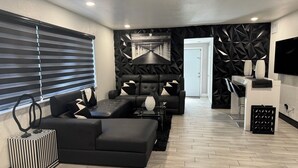 A modern living room with a black leather sofa, a black ottoman, and a black and white striped ottoman. There is a black and white patterned wall and a black geometric wall panel. A flat-screen TV is mounted on the wall, and a small table with a vase and a lamp is in front of the sofa. - MASSIVE HOME WITH HEATED POOL, NEAR BEACH IN SUNNY SOUTH FLORIDA  (Dania Beach)