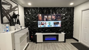 A modern living room with a flat-screen TV mounted on a geometric-patterned wall, a fireplace with a blue light, and a white entertainment unit. - MASSIVE HOME WITH HEATED POOL, NEAR BEACH IN SUNNY SOUTH FLORIDA  (Dania Beach)