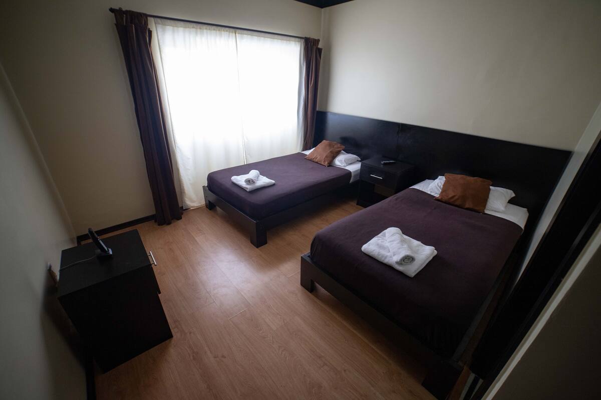 A hotel room with two beds, a bedside table, a nightstand, and a window with curtains.