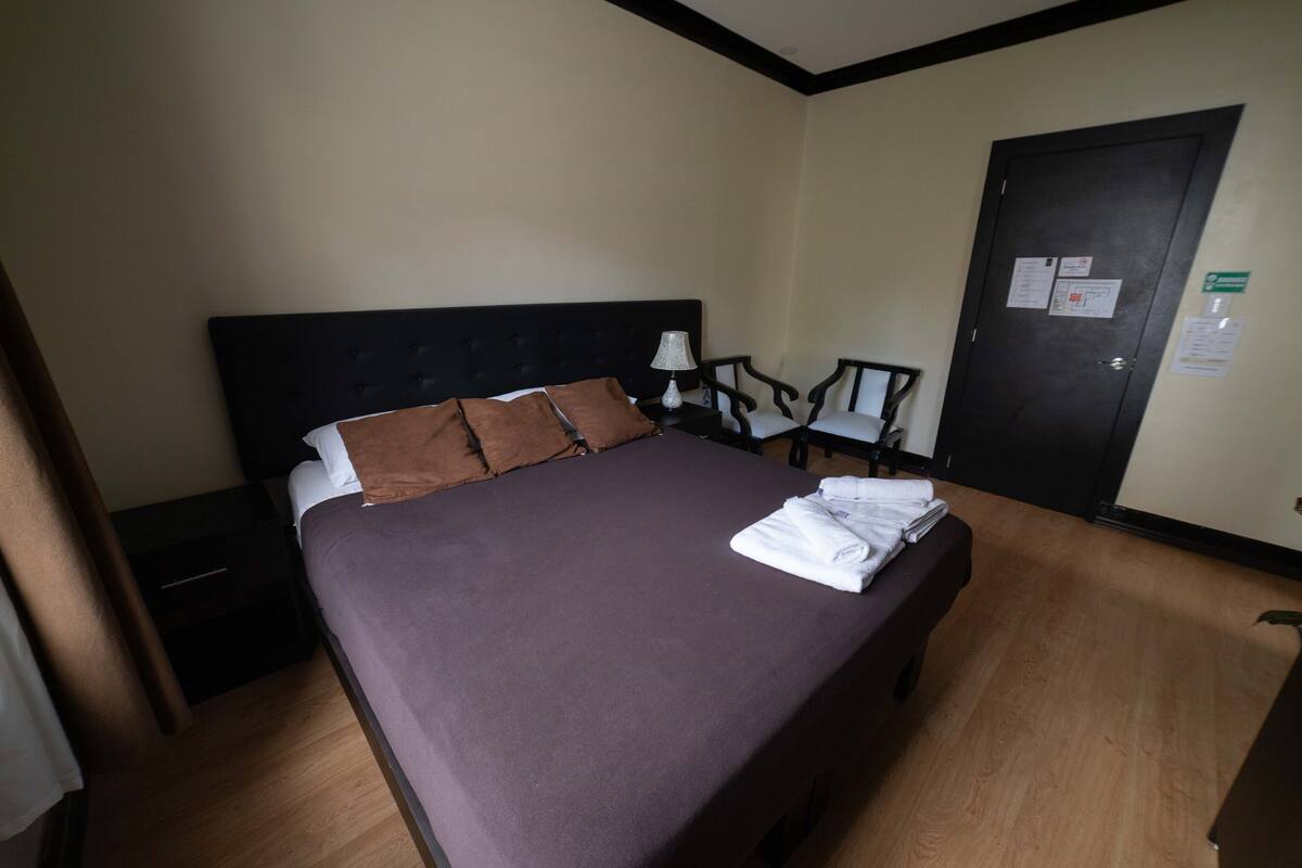 A hotel room with a large bed, a nightstand, a lamp, and a door with posted notices.