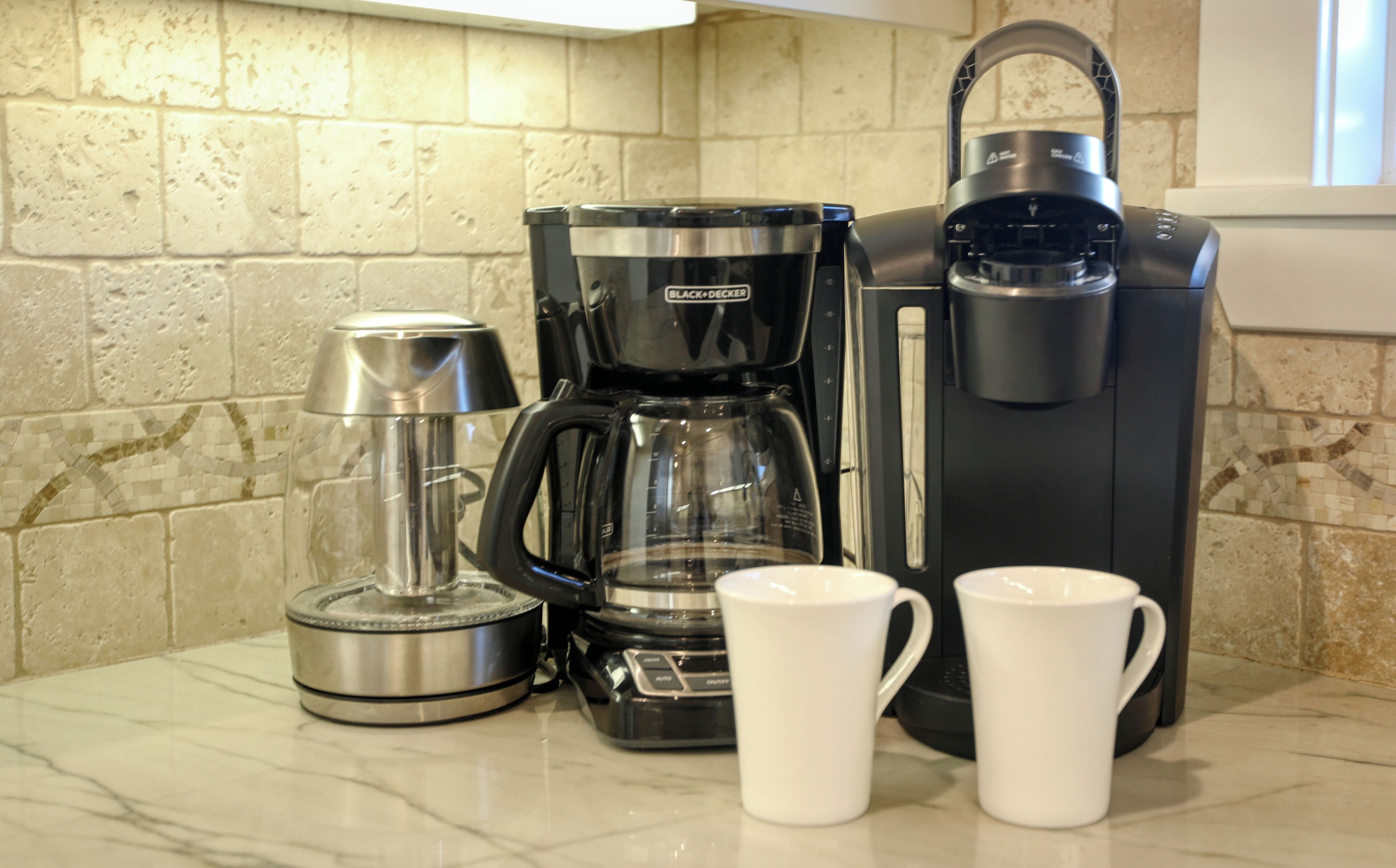 A kitchen with a coffee maker, a kettle, and two white mugs on a marble countertop.