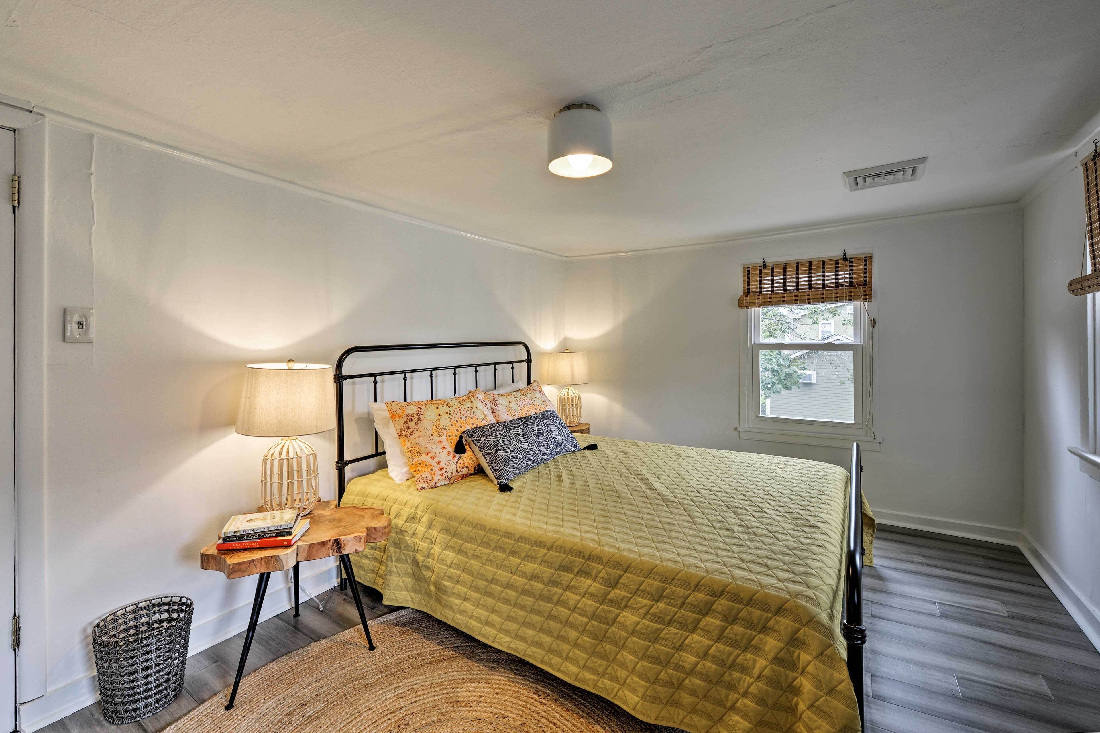 Ein Schlafzimmer mit einem Bett, Nachttischlampen, einem Fenster mit Jalousien, einem kleinen Tisch mit Büchern und einer Stehlampe.
