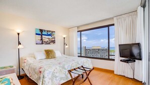 Une chambre d’hôtel avec un lit, une télévision, une grande fenêtre donnant sur des immeubles et l’océan, et un tableau accroché au mur.