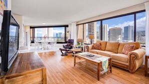 Un vaste salon doté d’une grande fenêtre offrant une vue sur la ville, d’une table basse en bois, d’un canapé beige agrémenté de coussins à motifs et d’un coin repas avec des chaises et une table blanches.