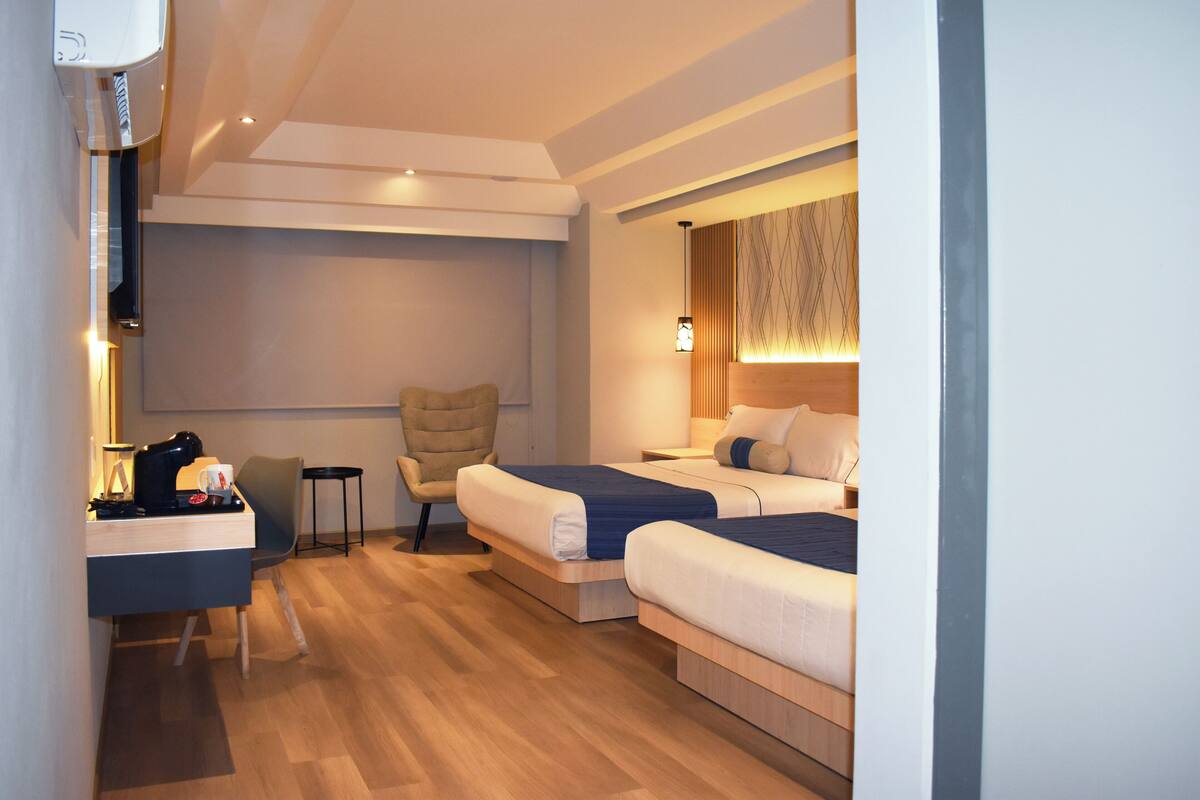 A modern hotel room with two single beds, a desk with a coffee maker, a chair, and a small table.