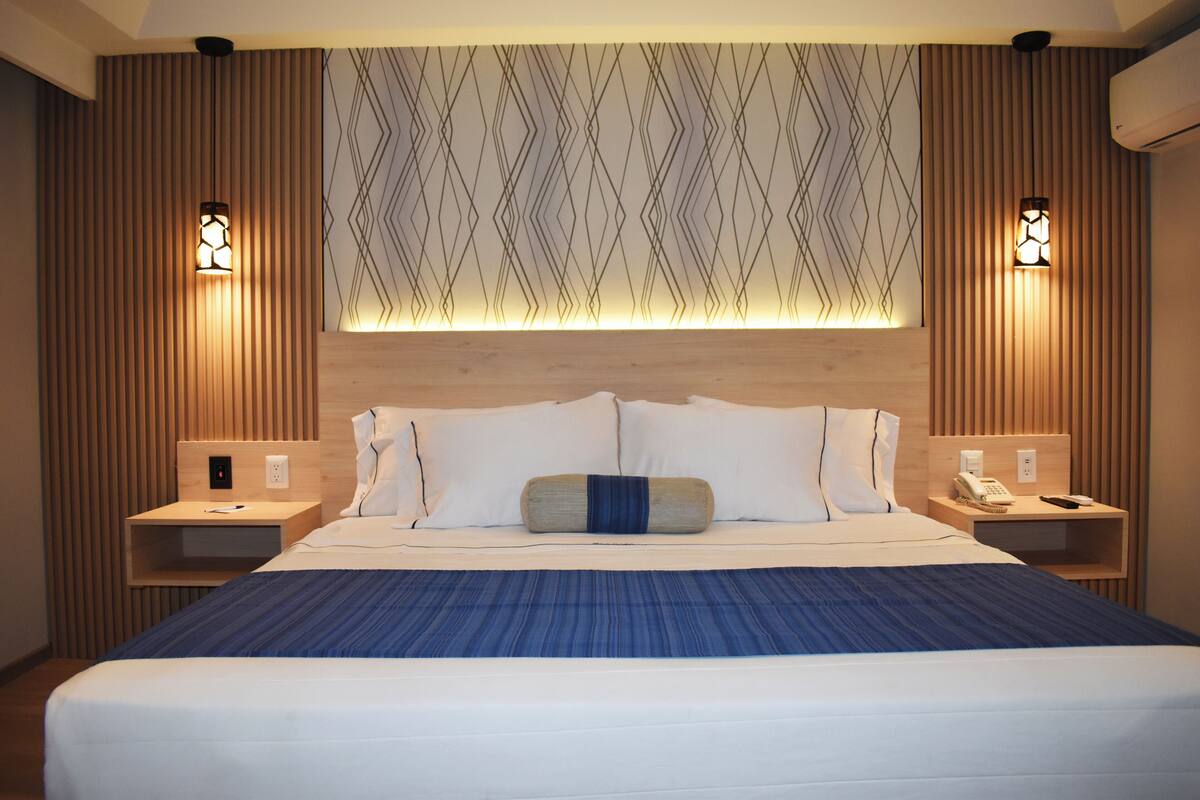 A hotel room with a large bed, wooden headboard, two bedside tables with lamps, and a patterned wall above the bed.