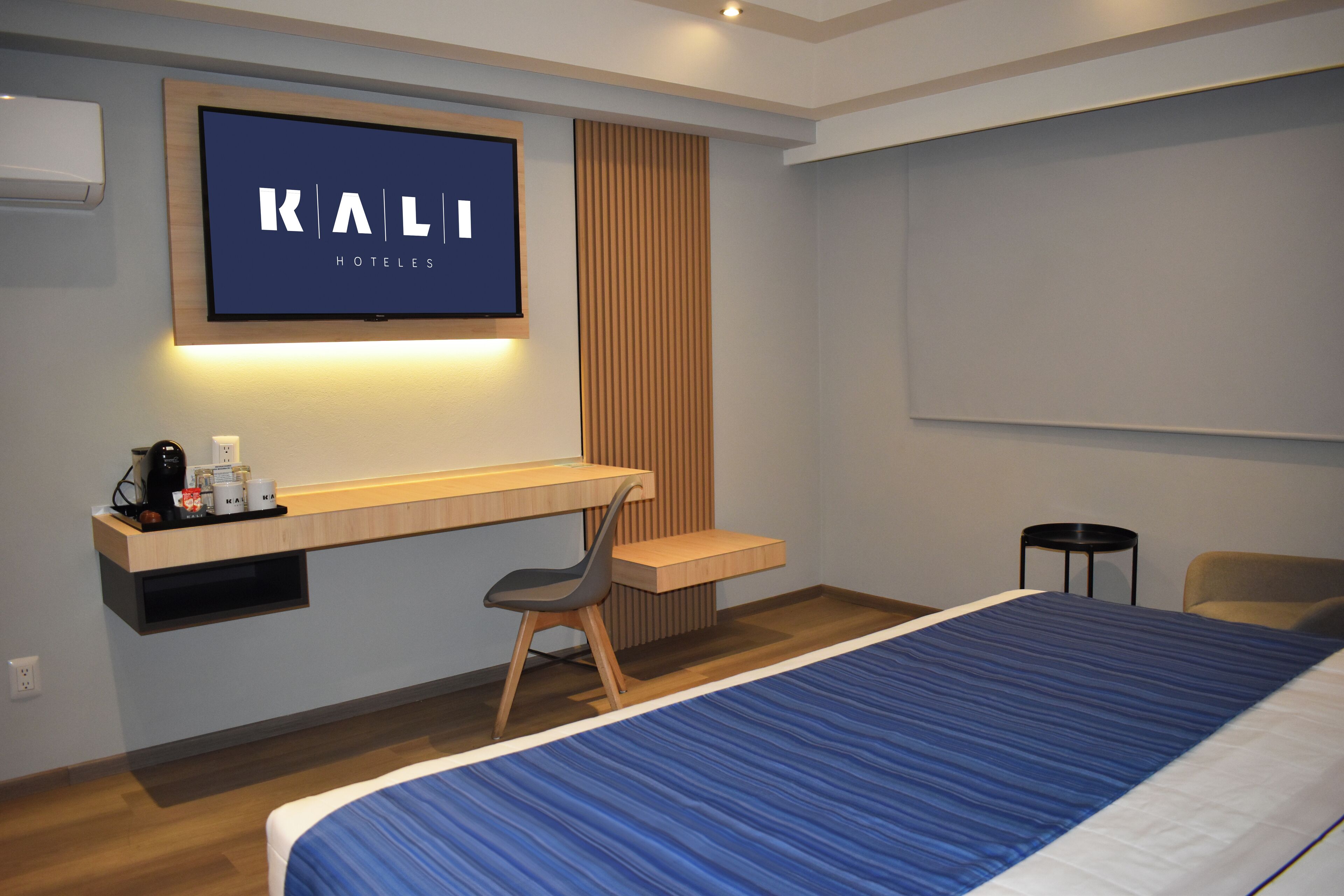 A modern hotel room with a bed, a desk, a chair, and a TV mounted on the wall displaying "KALI HOTELS".